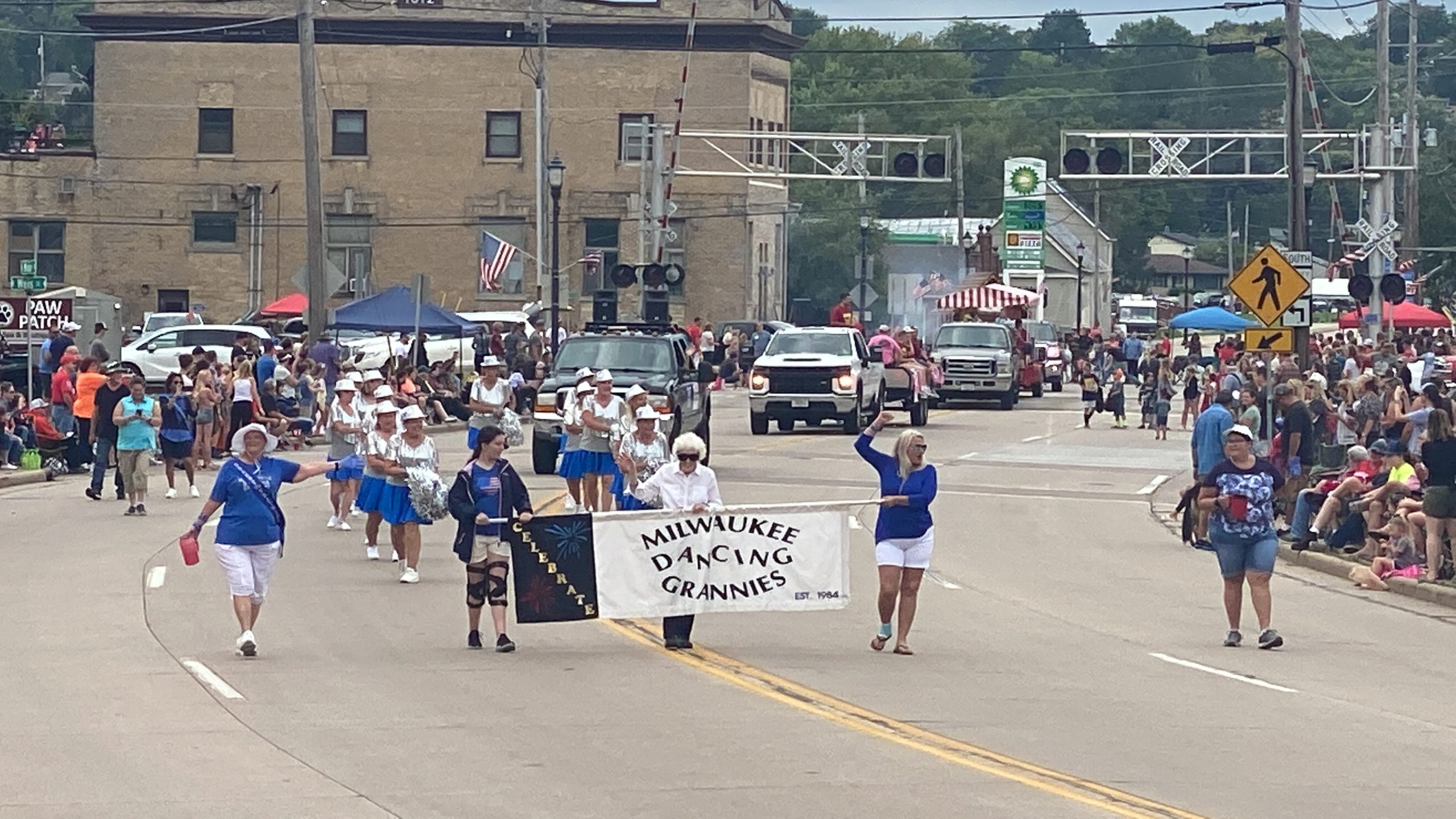 Two women hold a banner in the center of the image that reads "Milwaukee Dancing Grannies" with performers behind them. They hold pom poms and perform to music as part of the annual Allenton Parade.