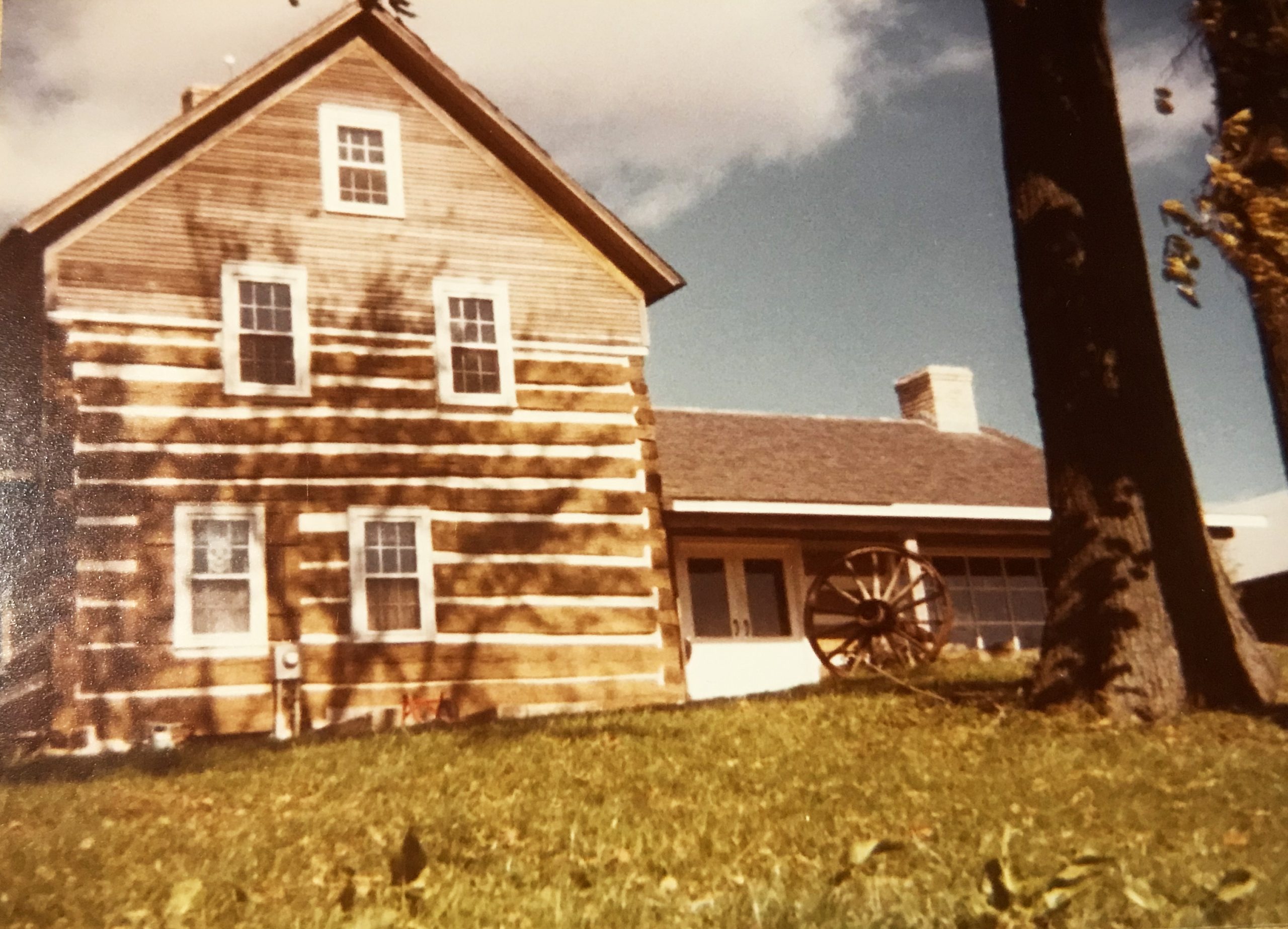 A two story log cabin with brown and white beams that has five windows and white double doors is pictured on a sunny day with some clouds that break up the blue sky.