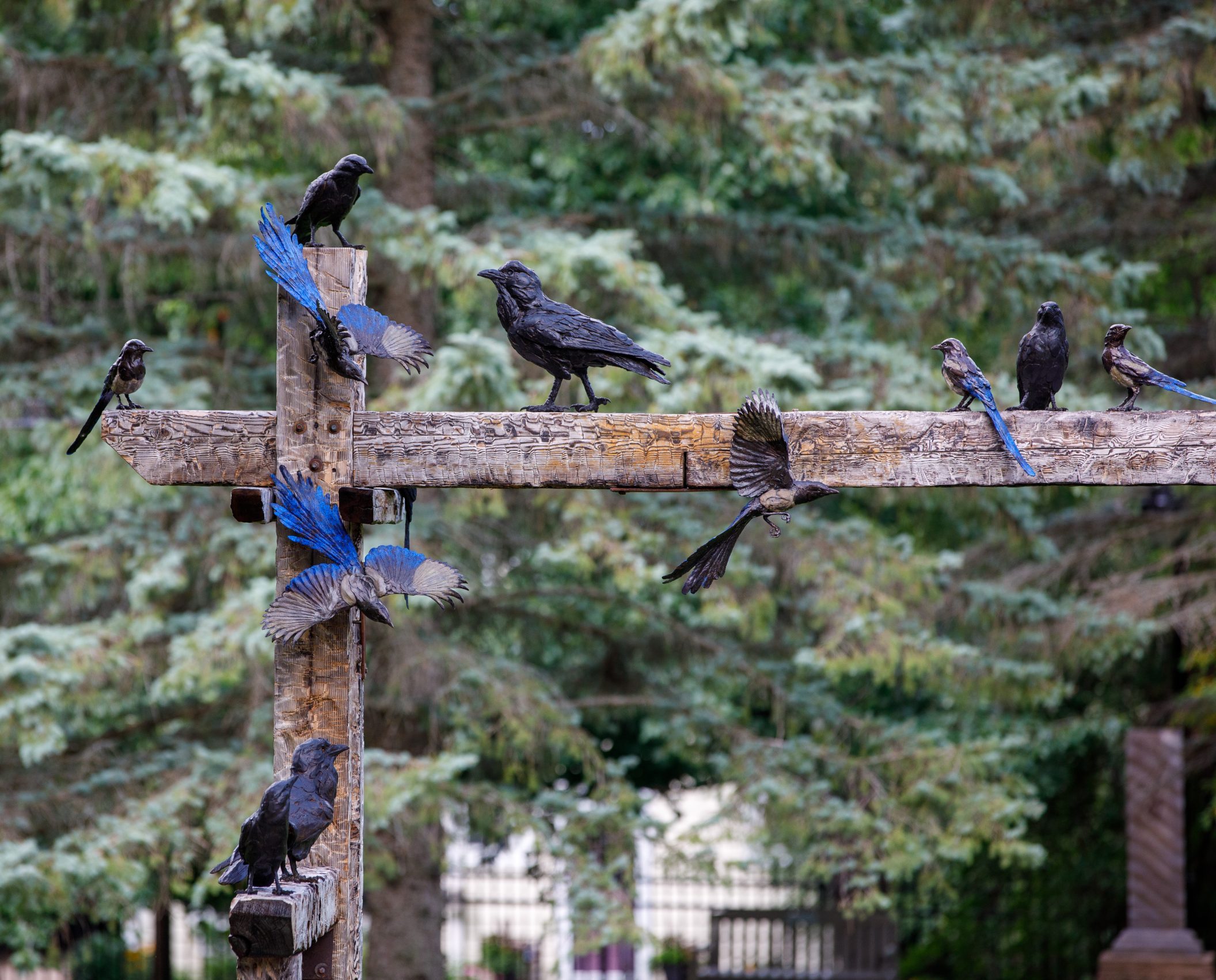 This image depicts a bronze and wood sculpture by artist Paul rhymer of a group of 19 crows, ravens, jays, and magpies interacting on wooden posts and beams