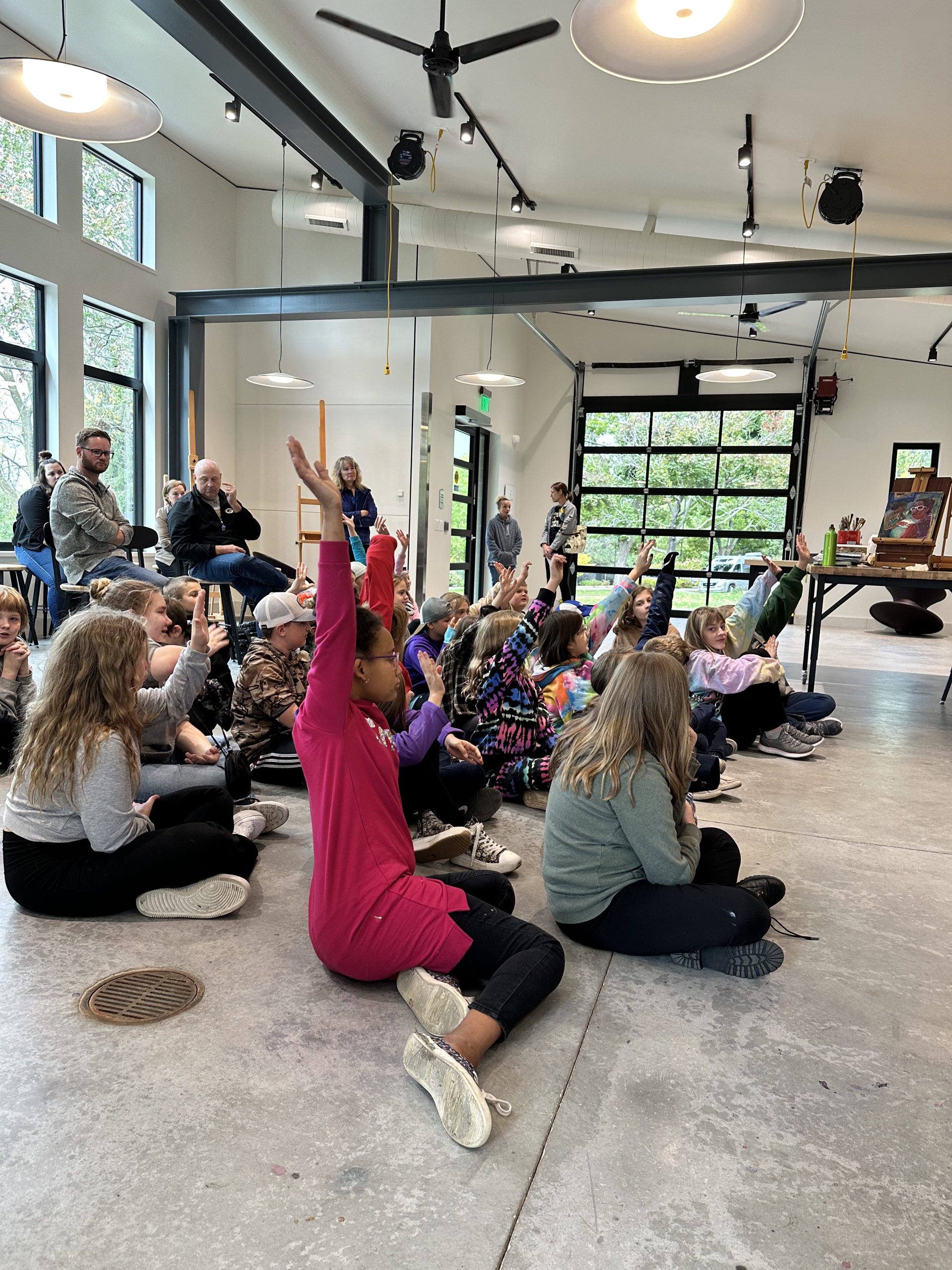Several students sit on the concrete floor of the Museum's Glass Box Studio. They are engaged and raising hands to ask questions of the artist.