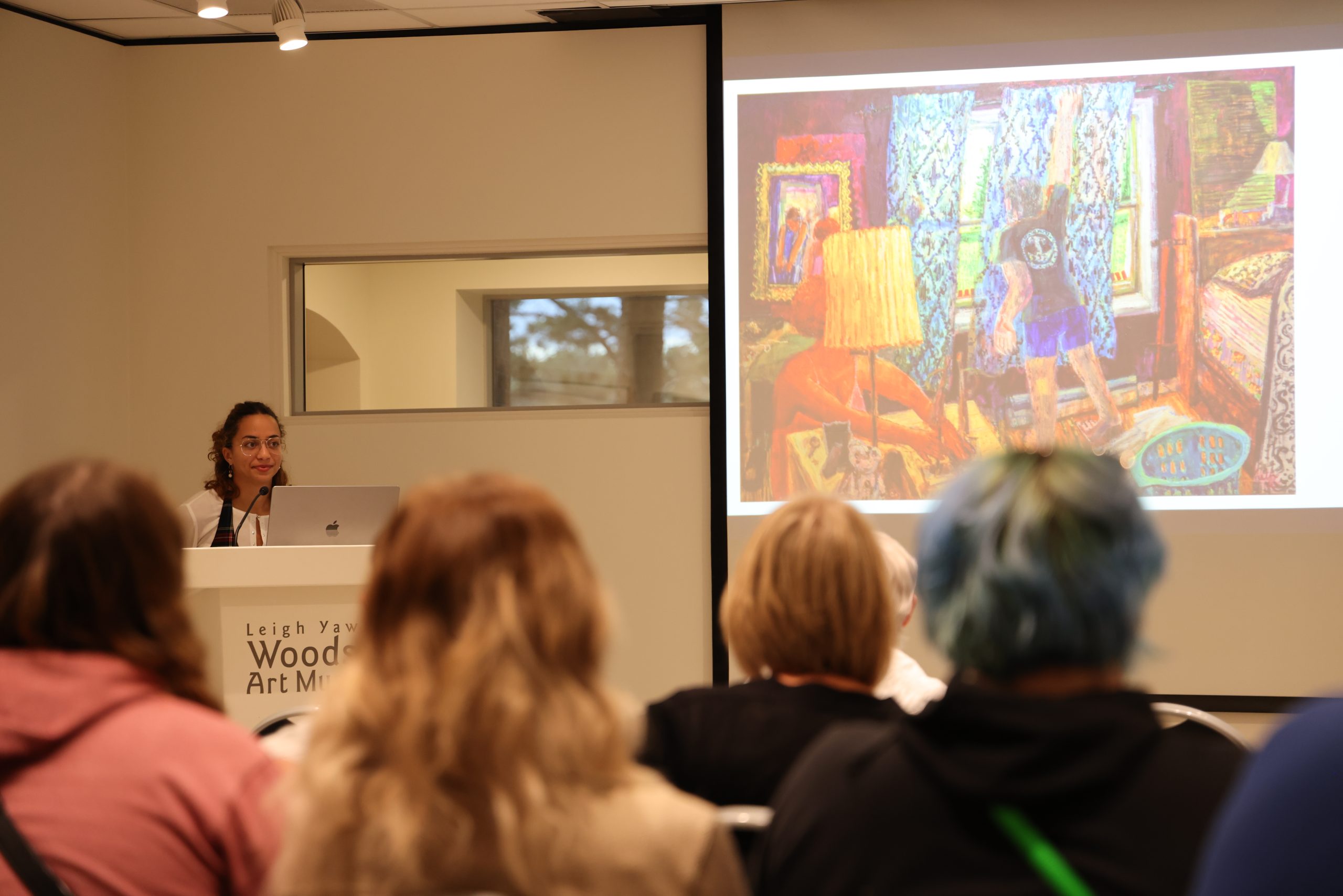 Ariana Vaeth stands behind a podium to the left of a projector screen. She is giving an artist talk about her work in the Museum's classroom space. The screen depicts a painting of hers with lots of colorful patterns and a figure reaching upwards. 