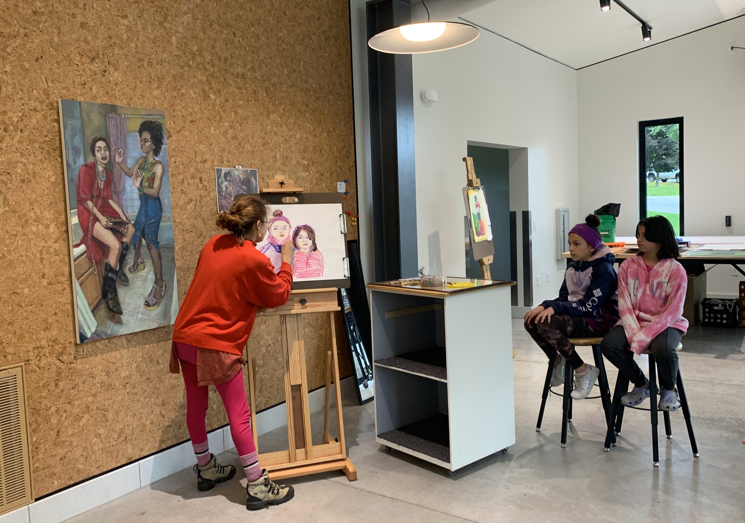 Two students sit on stools on the right side of the image. Beside them, is Ariana Vaeth, October Artist in Residence, dressed in all red. She is creating a drawing of the students using oil pastels and an easel in the Museum's Glass Box Studio. The drawing is made up of pinks, purples, and flesh tones.