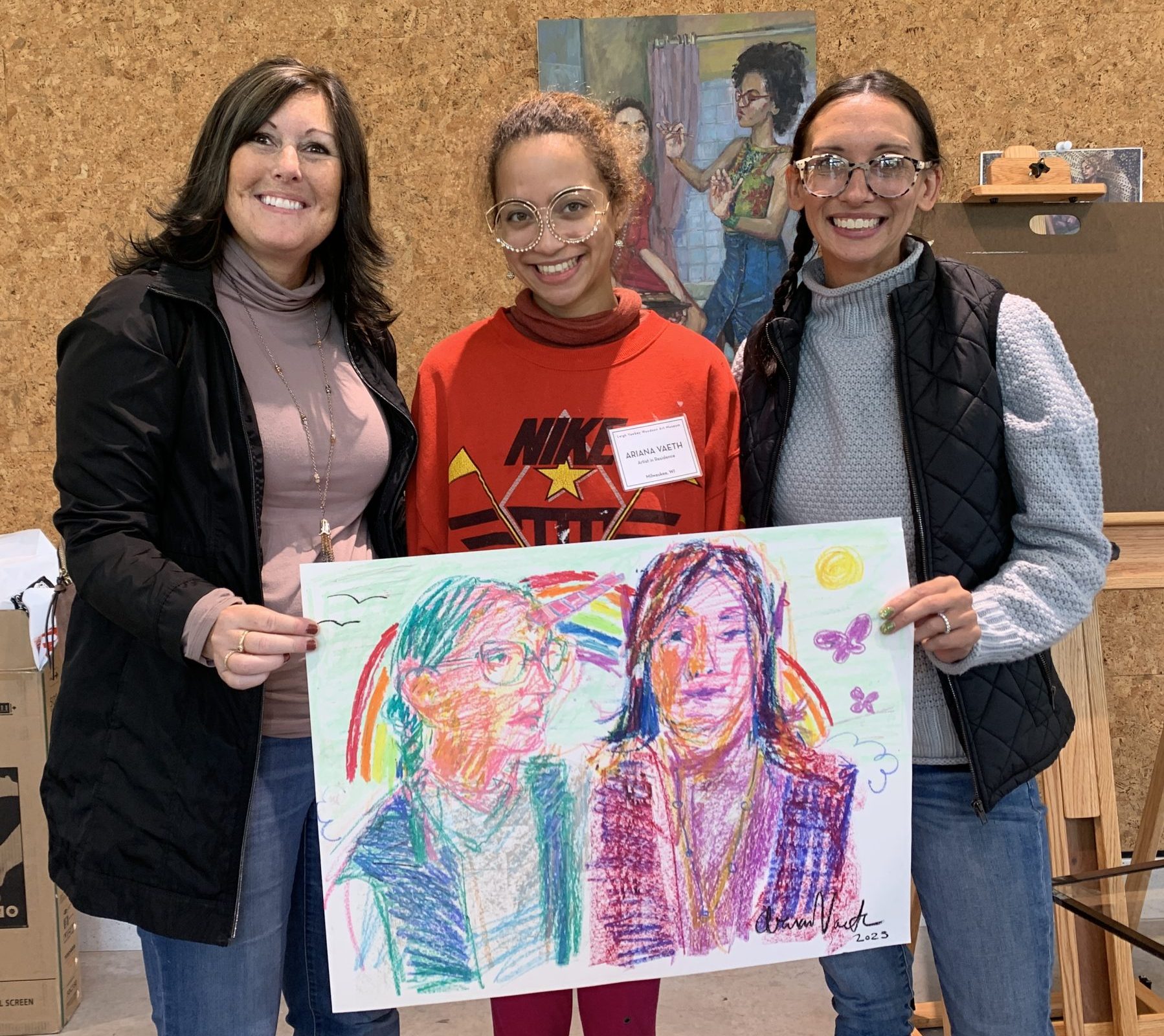 Three women stand together holding a colorful drawing created by Ariana Vaeth. The drawing was created in oil pastel with lots of purples, pinks, and blues. All three people are smiling happily. 