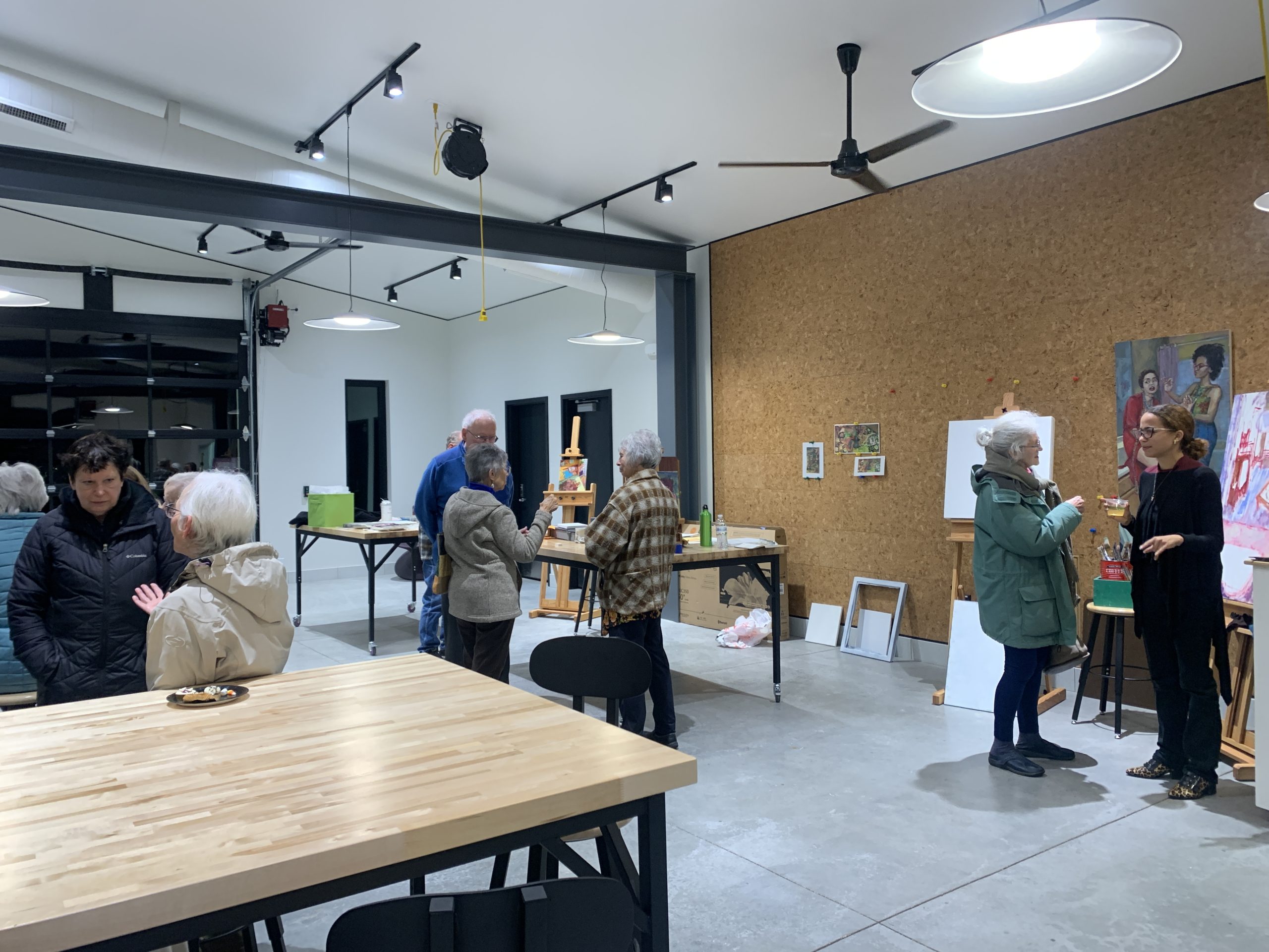 People are mingling in the Glass Box Studio, a large studio space with concrete floors and industrial beams and wood topped tables. Artist in Residence Ariana Vaeth stands on the right side of the frame talking to a Bird Club participant. A cork wall is behind them with paintings and drawings hanging up on it. 