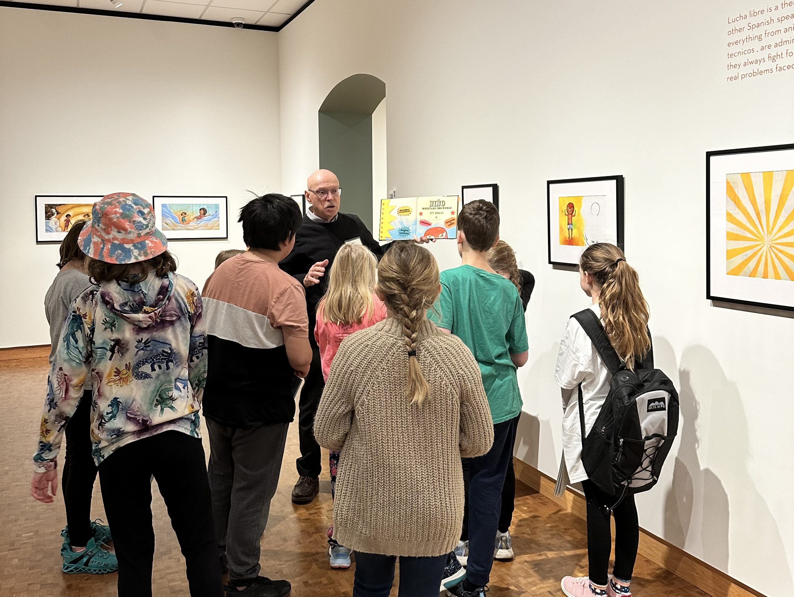 Students stand in a large gallery space with colorful mixed media illustrations. They are facing a museum docent, who is holding up a book illustrated by artist Yuyi Morales