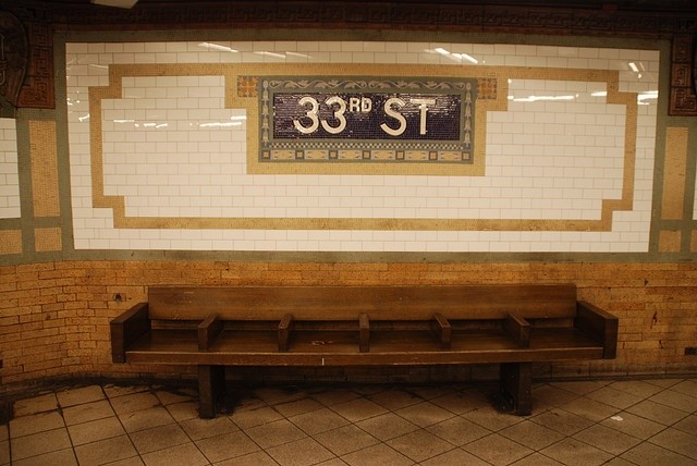 Image of a long bench with center deviders with subway tiles on the wall behinds that say 33rd st