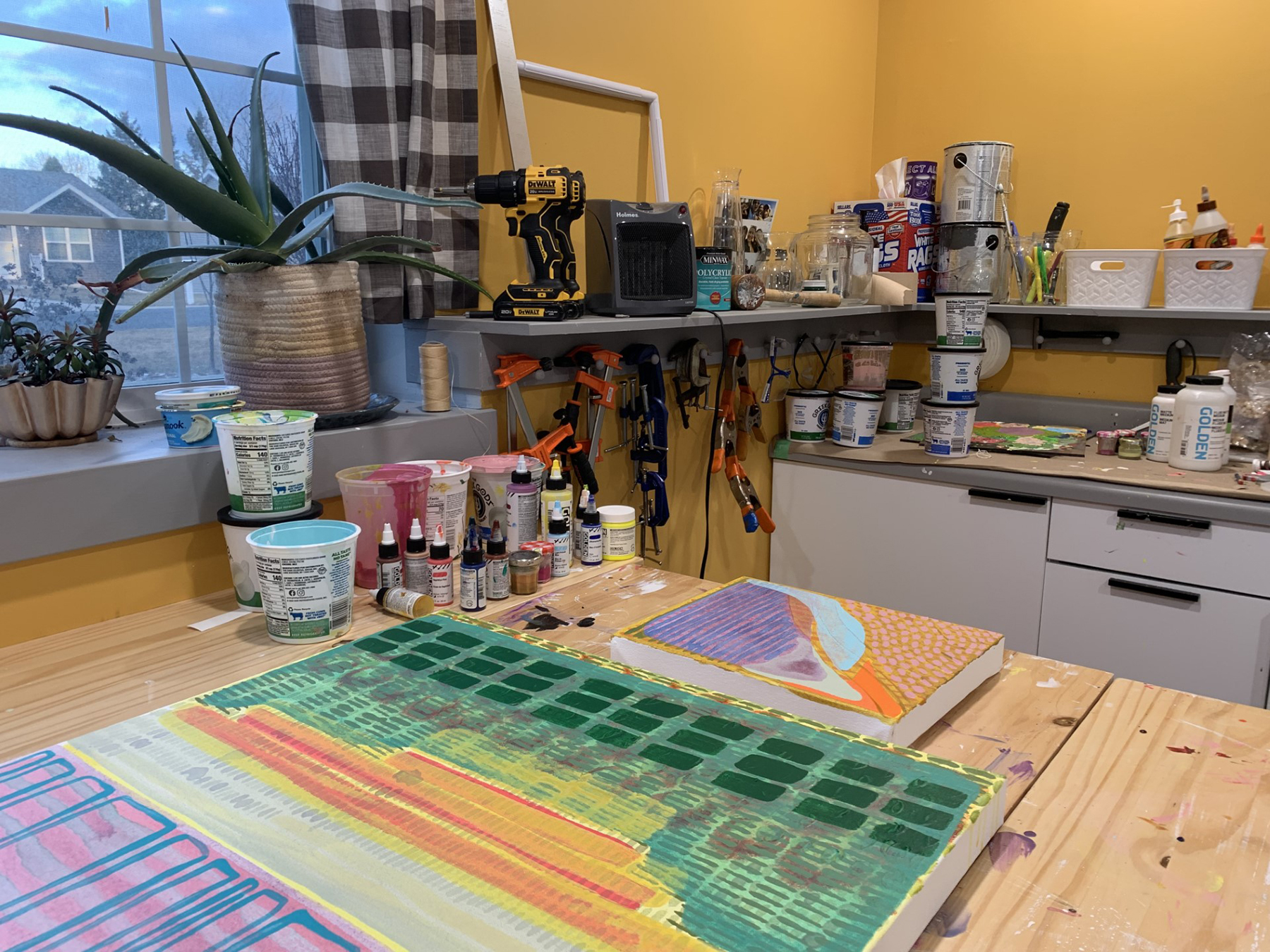 Photograph of an artist studio, paintings in the forground with paints and supplies behind.