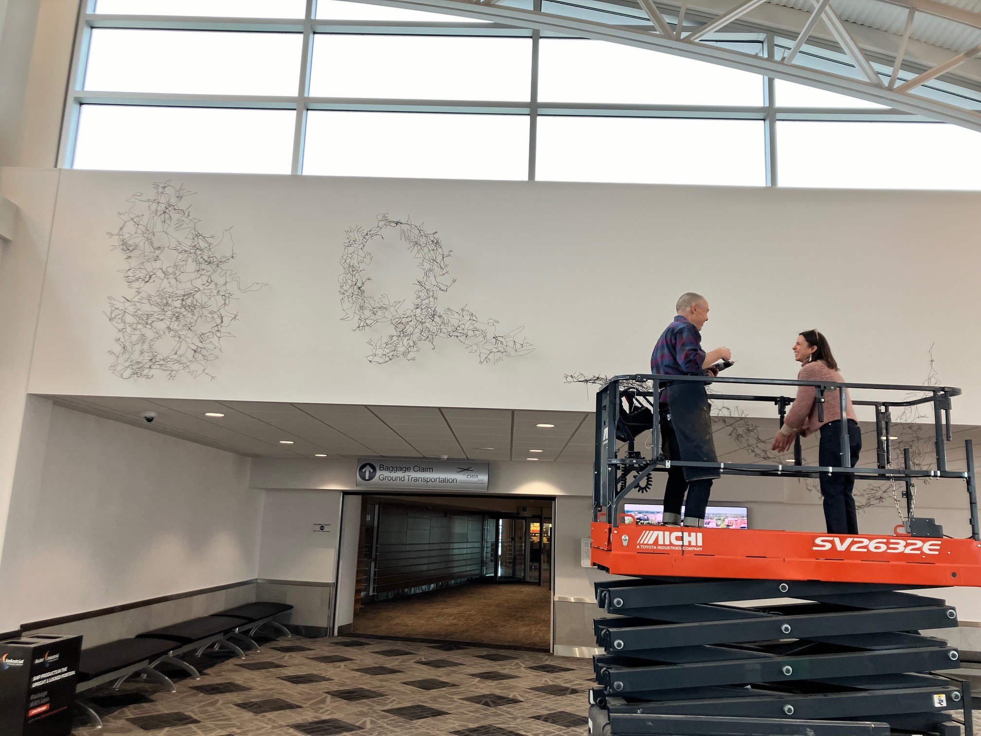 This image shows artist Tom Hill on a large lift with Museum curator of exhibitions Shannon Pueschner installing "Flock Together"