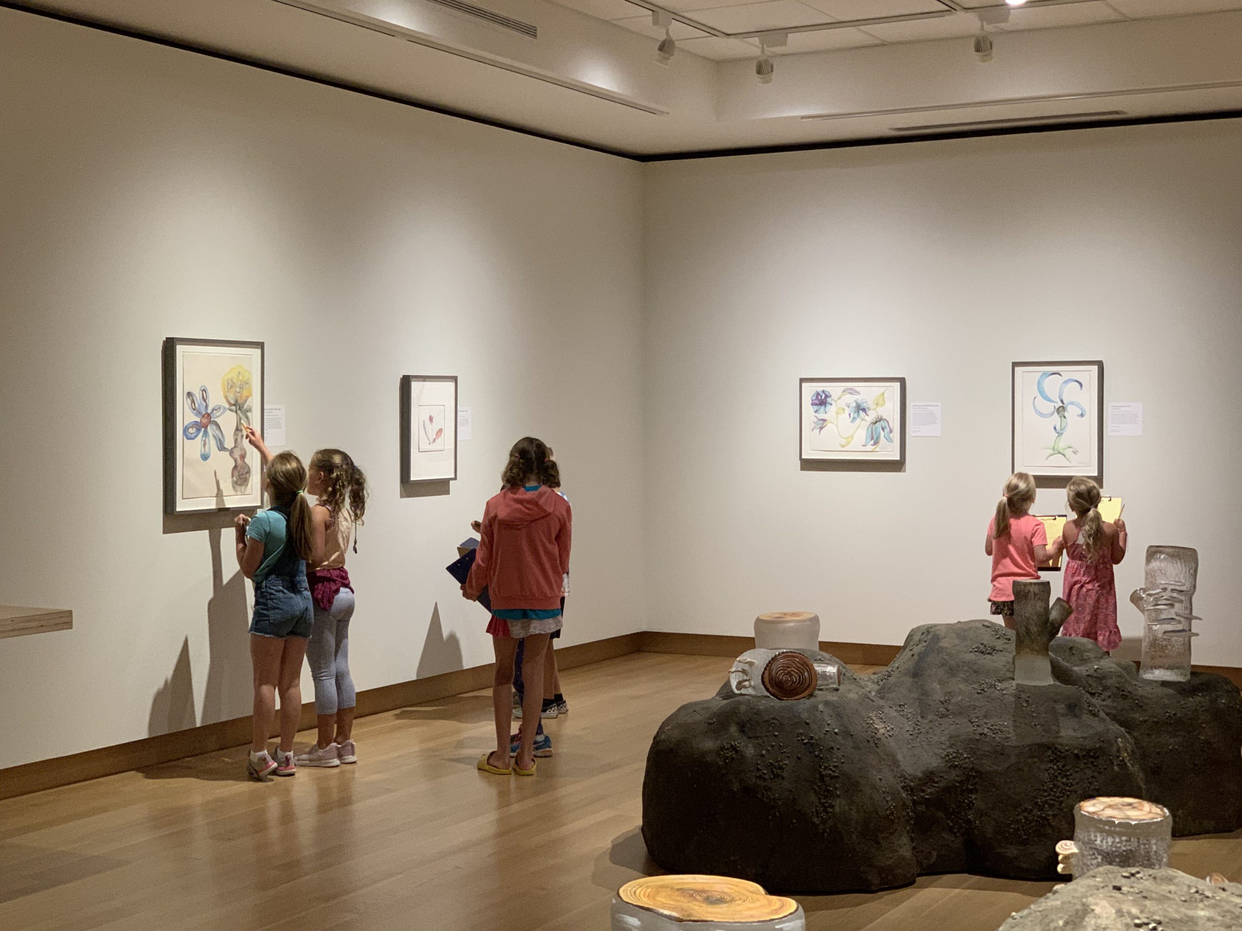 Three pairs of Summer Art Session participants, holding clipboards, look up at Ginny Ruffner paintings in a corner of her "Reforestation of the Imagination" gallery.