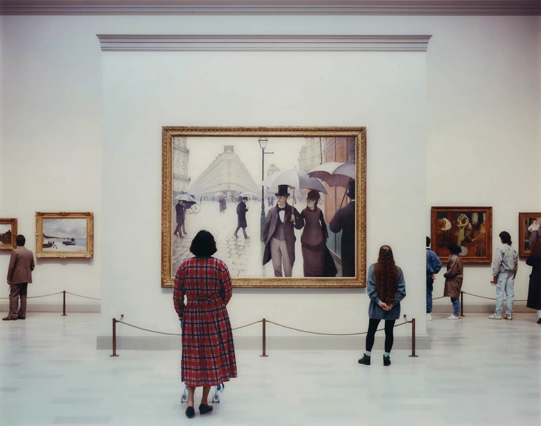 A woman in a red plaid dress stands in front of a very large painting of two walkers in black under an umbrella in Paris.