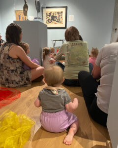 Babies and adults sitting on the floor in a gallery looking at the artworks.