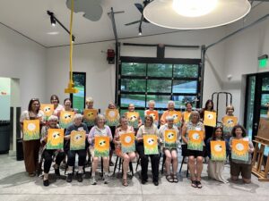 a group of volunteers standing in the glass box studio holding their canvases they painted on during the party.