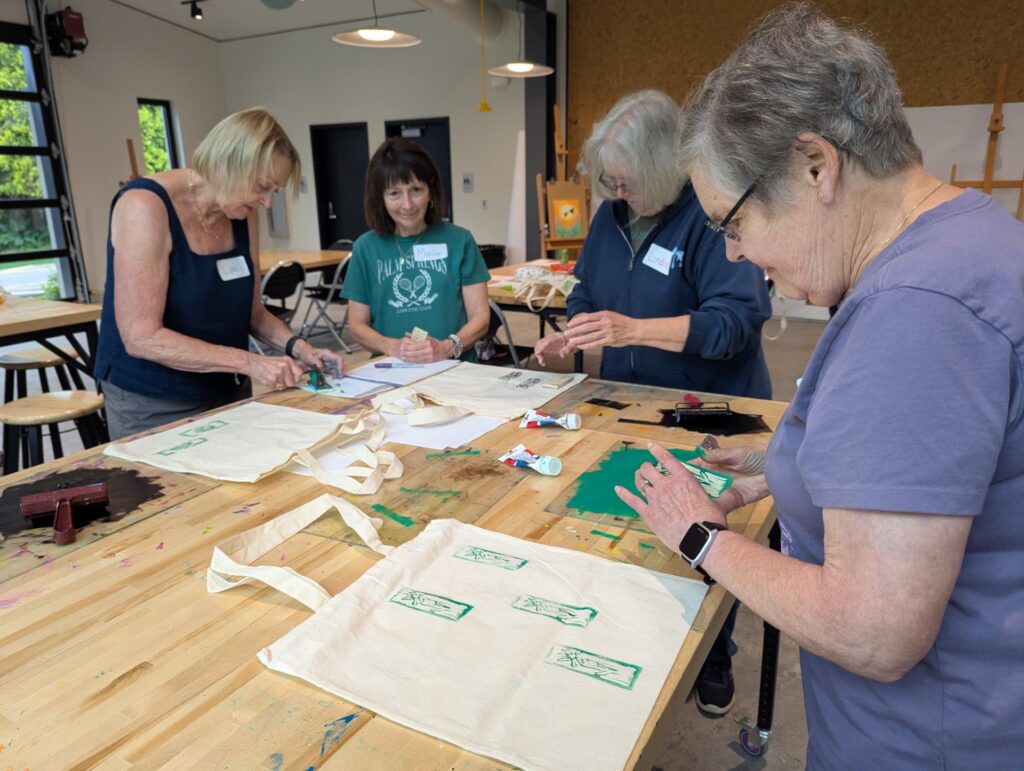 four ladies standing at a table in the glass box studio working on their handmade linocut prints during their party.