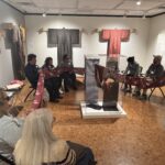 Several adults sitting in a circle in a gallery space holding onto a piece of cloth that spans the entire circle.