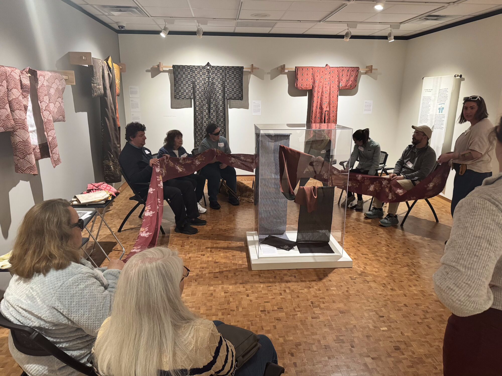 Several adults sitting in a circle in a gallery space holding onto a piece of cloth that spans the entire circle.