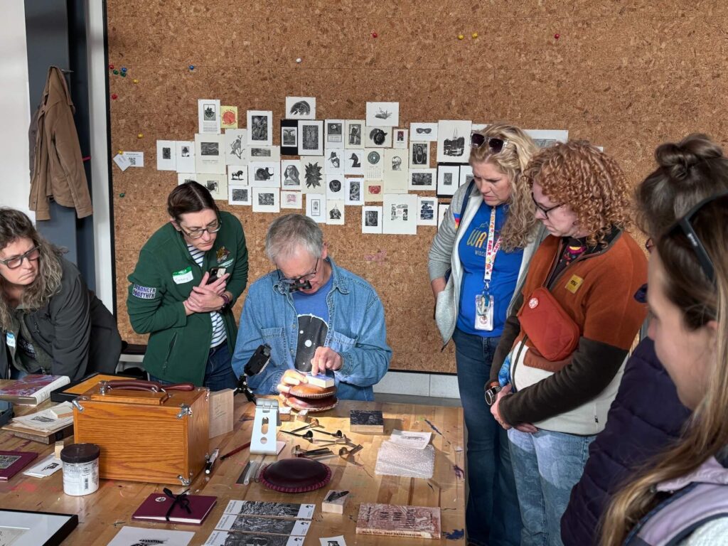 Several Wausau School District art educators gathered around Tony at a table, observing while he works on a wood engraving