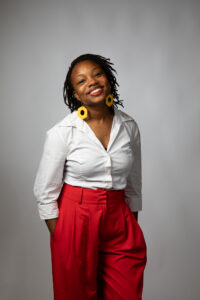 Photo credit: Adrienne Maples Woman stands in white shirt, red pants, and yellow flower earings smiles.